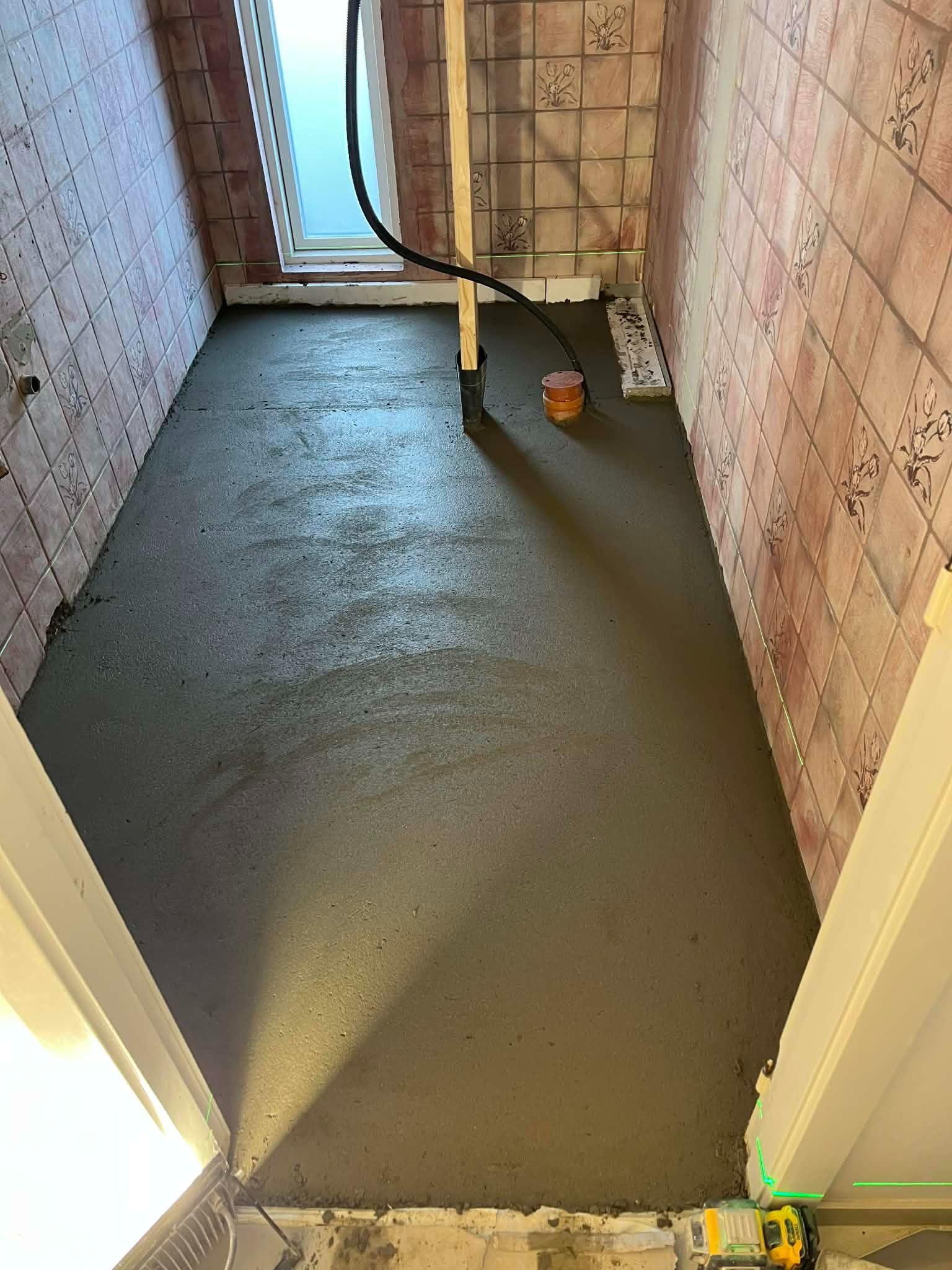 Ny betonbelægning i et badeværelse med fliser på væggene og en afløbsrør i midten.