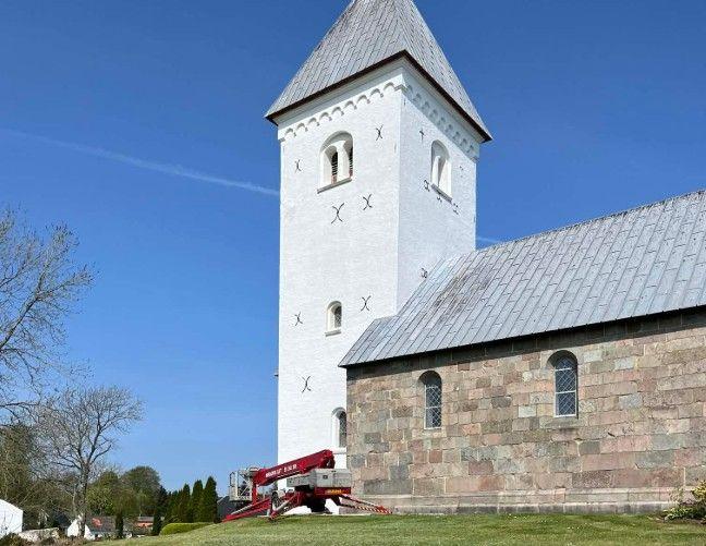 Hvid kirke med tårn og grå tag, bygget af sten, med en lift ved siden af til reparationer.