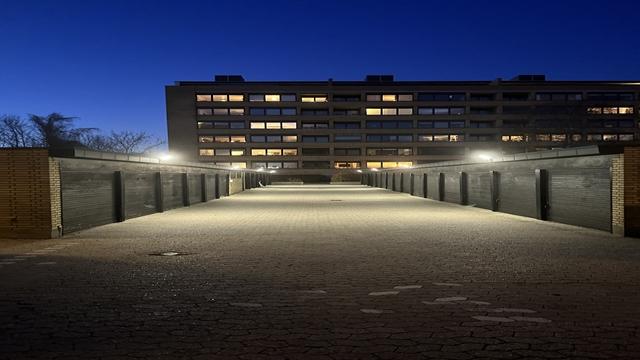 En belyst gangbro ved en bygning om natten, med mørk himmel og vinduer, der lyser op i baggrunden.