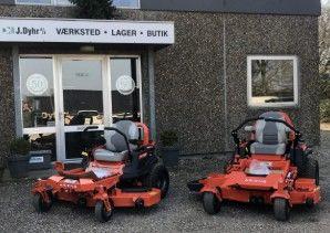 To store med to orange plæneklippere foran en bygning med skiltet "J. Dyhr" og glasdøre.