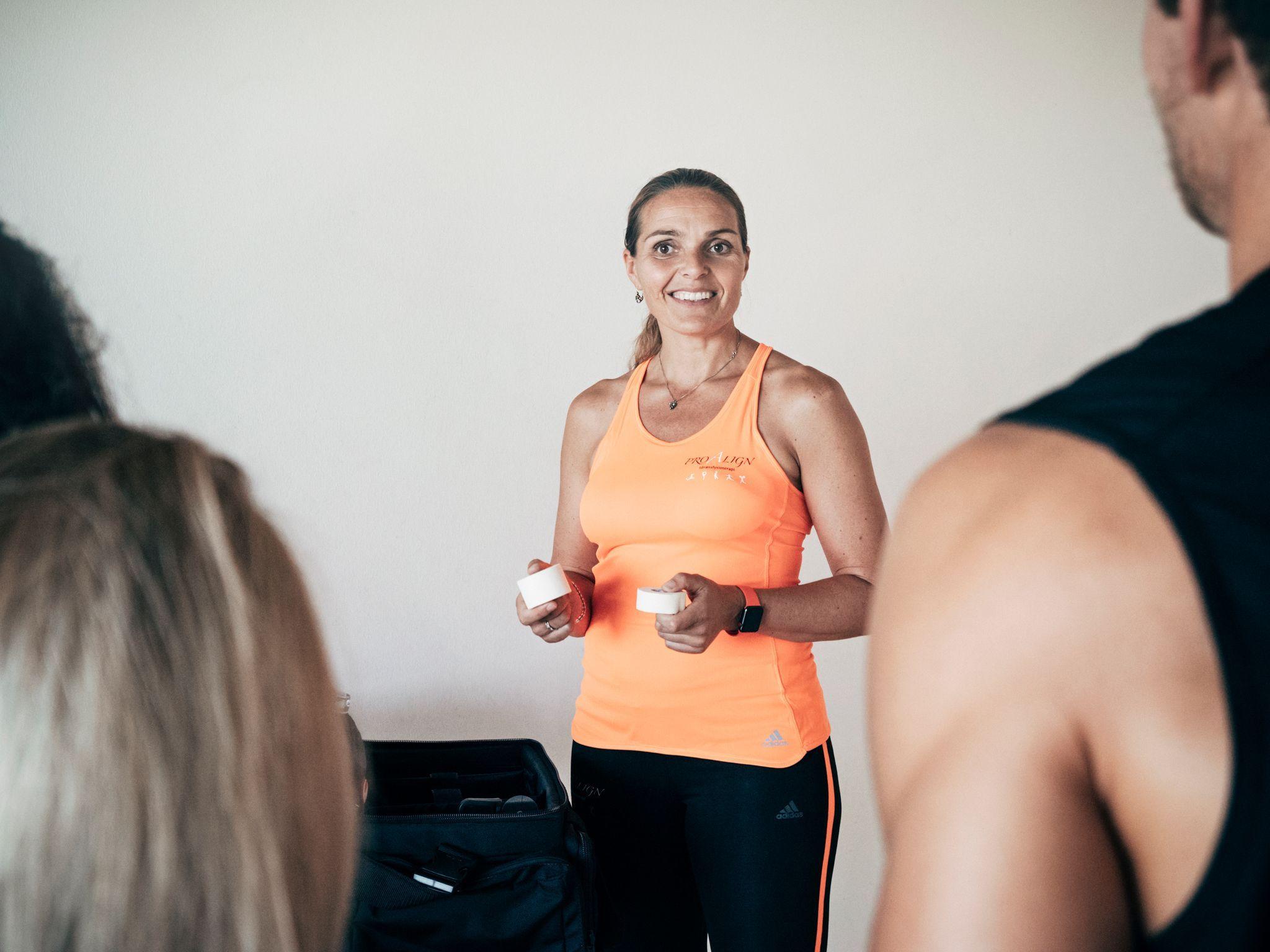 Træner i orange top, der holder to bolde, mens hun smiler til en gruppe mennesker.
