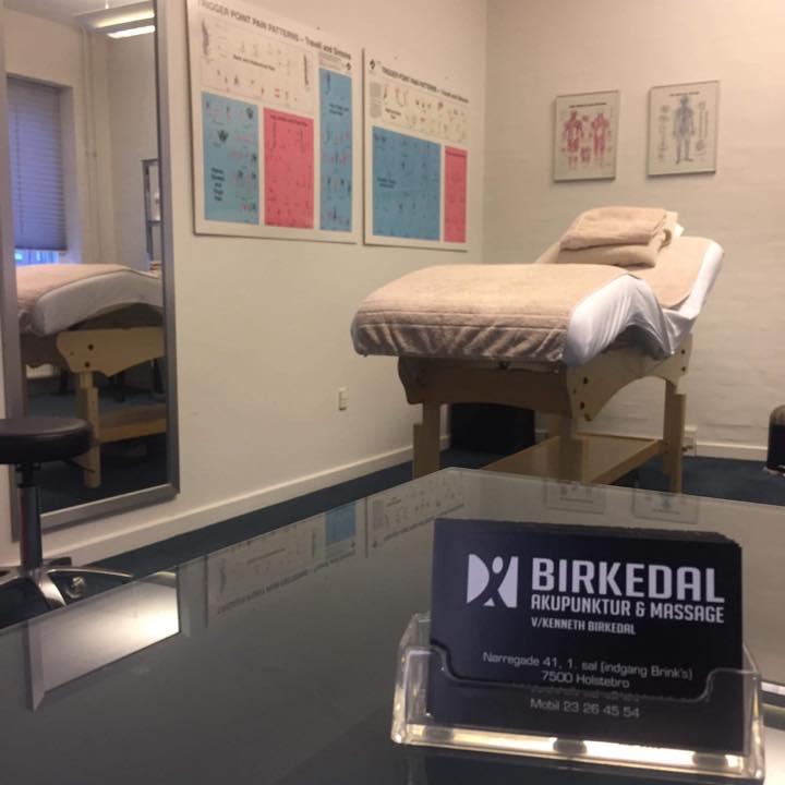 Behandlingsrum med massagebriks, akupunkturplakater på væggen og et skilt med navnet Birkedal Akupunktur & Massage.