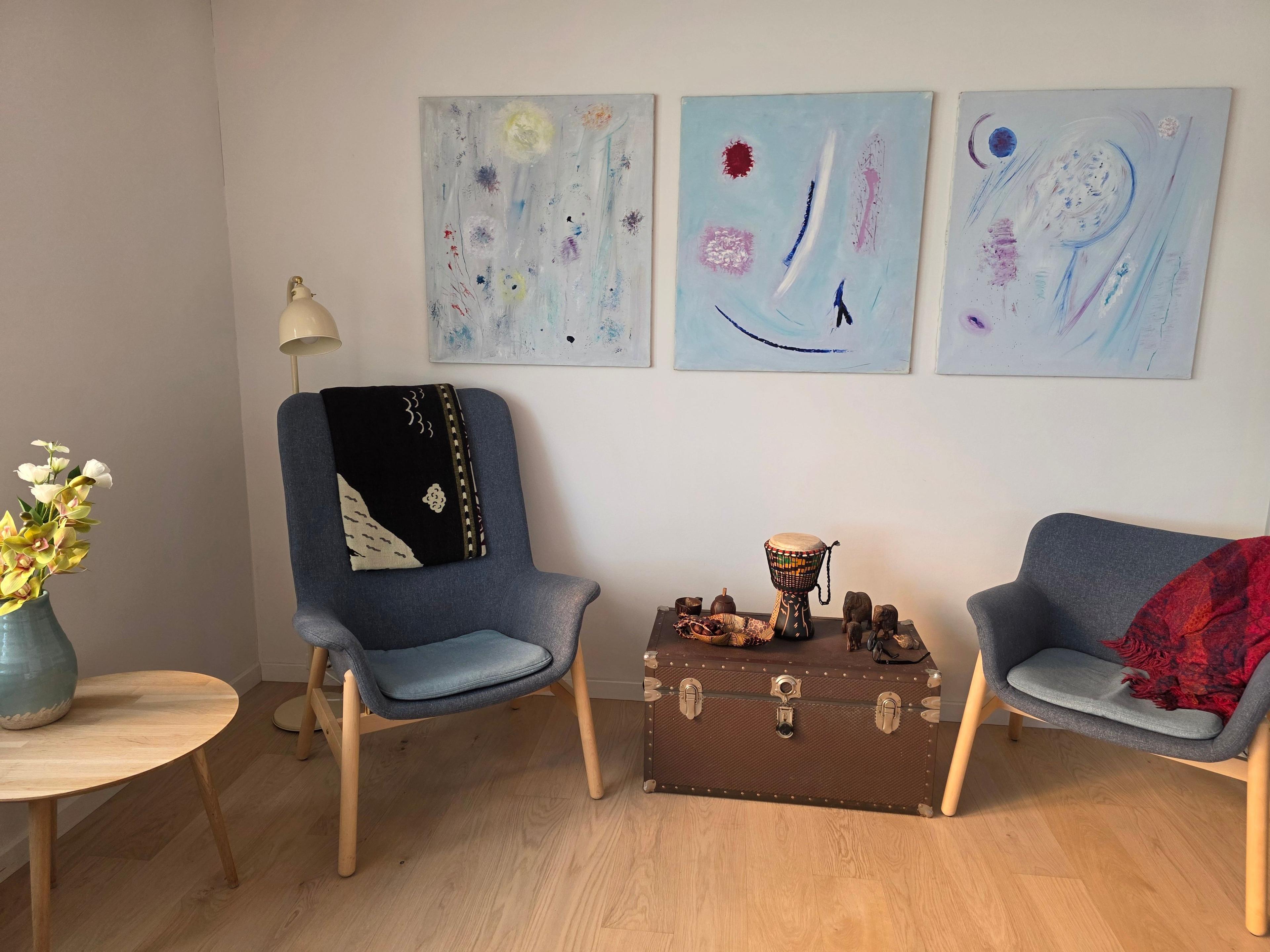 Hyggelig stue med to blå stole, et rundt bord, en lampe og tre farverige malerier på væggen.