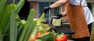 En person griller grøntsager som asparges og jordbær på en terrasse med en grill i baggrunden.