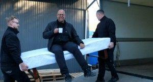 Tre mænd bærer en stor madras, mens en af dem sidder på den og holder en kop.