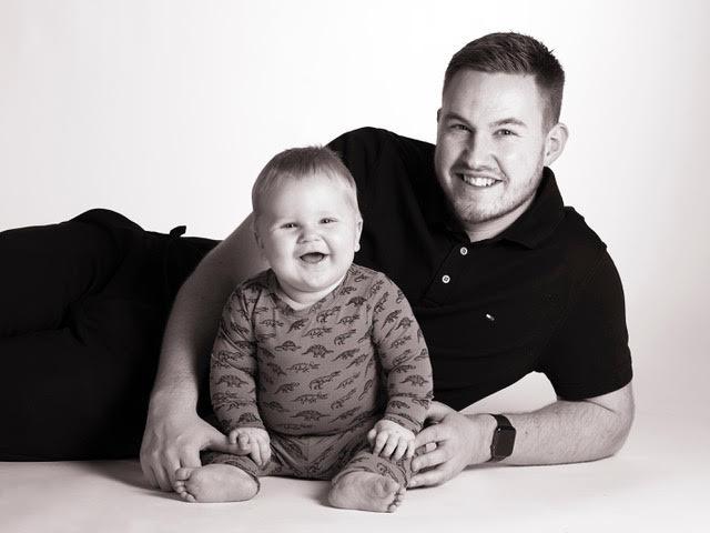 Far og baby ligger på gulvet og smiler til kameraet i en sort-hvid fotografering.