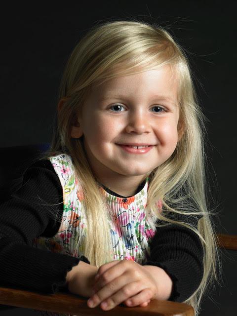 Lille pige med langt, lyst hår og smilende ansigt, iført en farverig bluse, sidder på en stol.