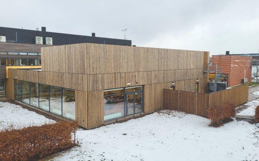 Moderne bygning med træfacade og store vinduer, omgivet af sne og buske.