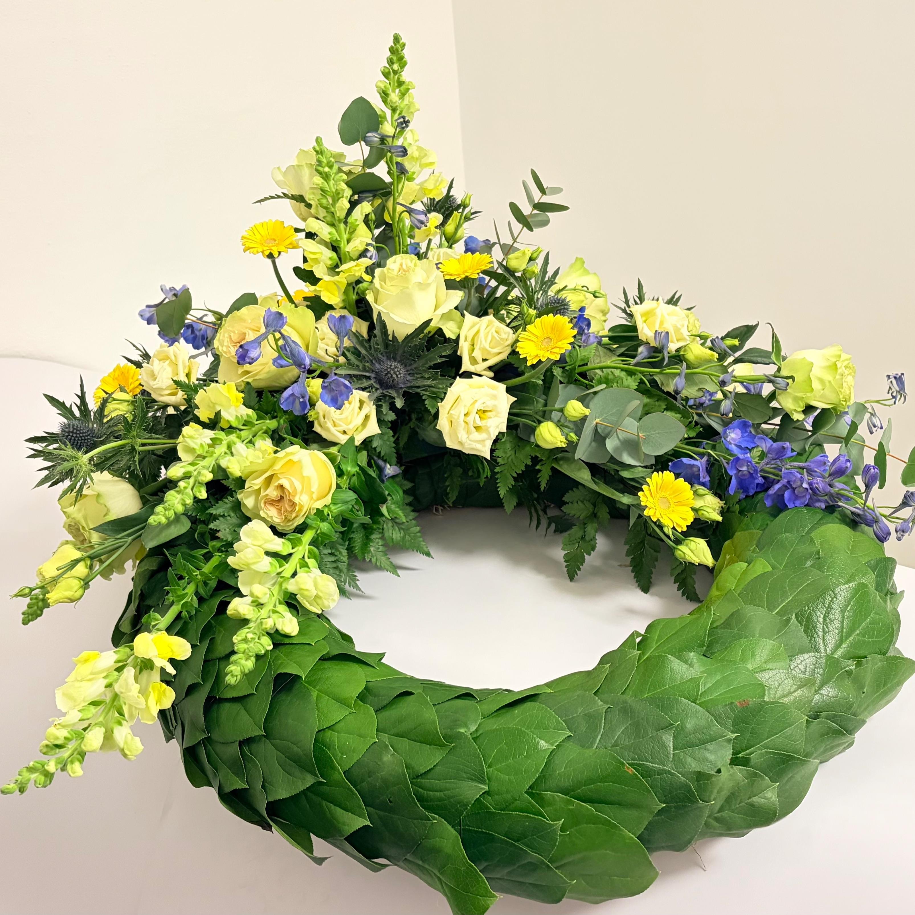 En smuk blomsterkrans med gule og blå blomster samt grønne blade, præsenteret på en hvid baggrund.
