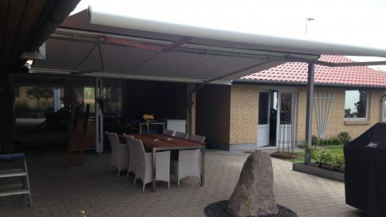 Terrasse med overdækning, spisebord og stole, samt en grill og en stor sten i haven.