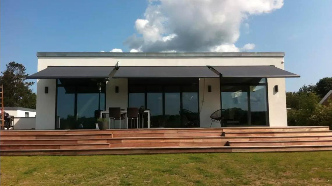 Moderne hus med store vinduer, terrasse og markiser, beliggende i et grønt område under en blå himmel.