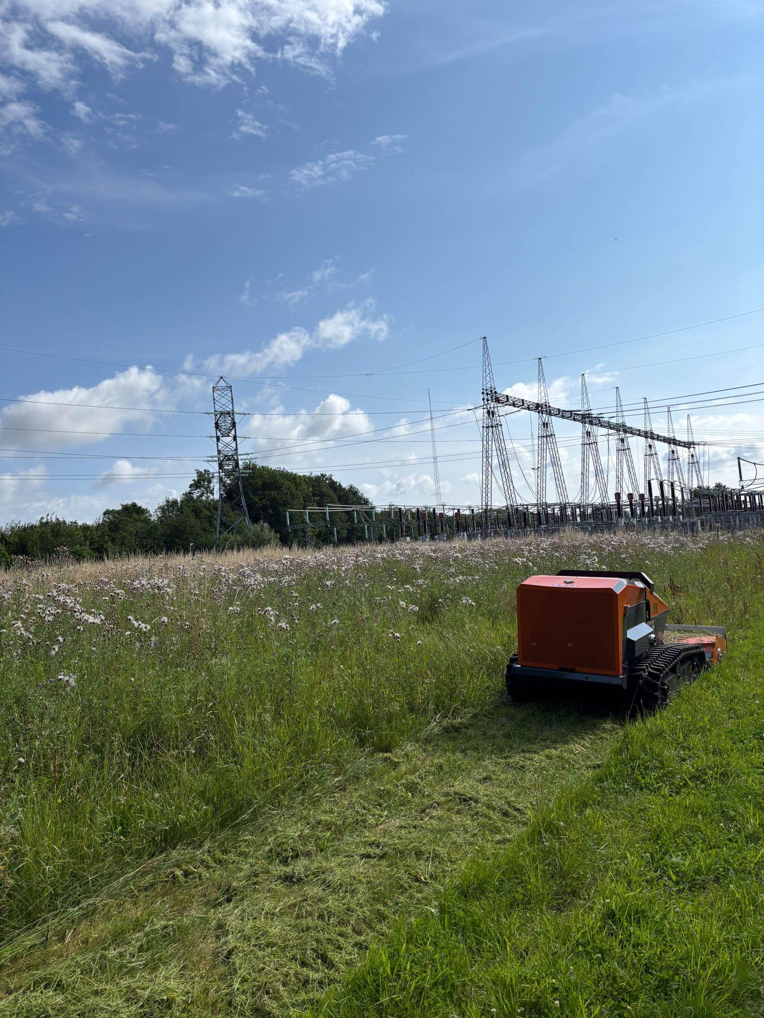 En græsslåmaskine klipper græsset i en mark nær en elektrisk station under en blå himmel med skyer.