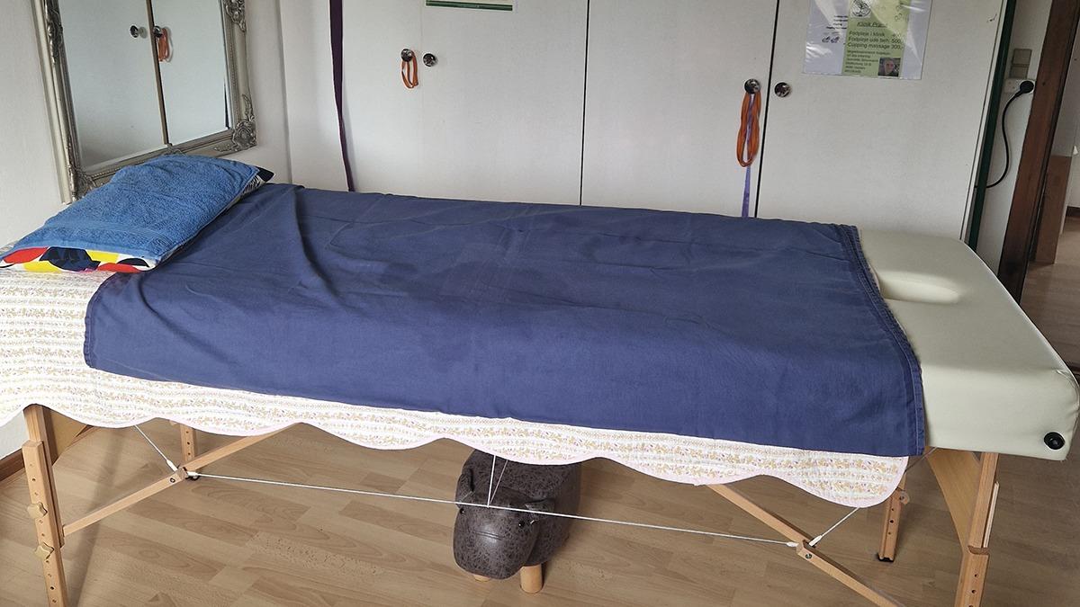 En massagebriks med en blå dug og en farverig pude i et lyst rum med skabe i baggrunden.