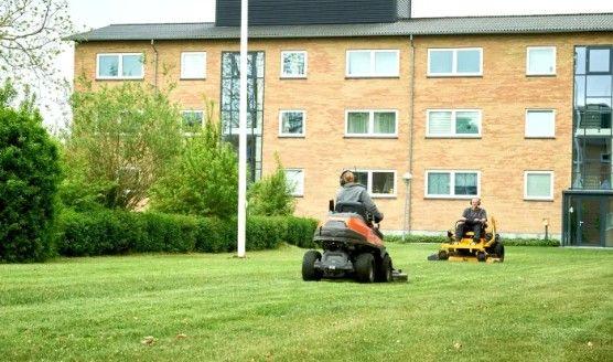 To personer klipper græs med plæneklippere i en grøn have foran en bygning med murstensvæg.
