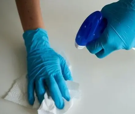 Hånd med blå gummihandsker, der sprayer rengøringsmiddel på en overflade, mens der tørres med et papirhåndklæde.