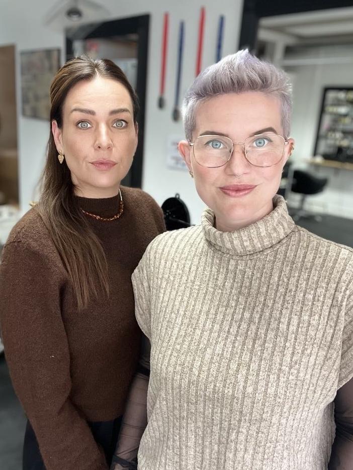 To kvinder står sammen i en frisørsalon, iført strikkede trøjer i brune og beige nuancer.