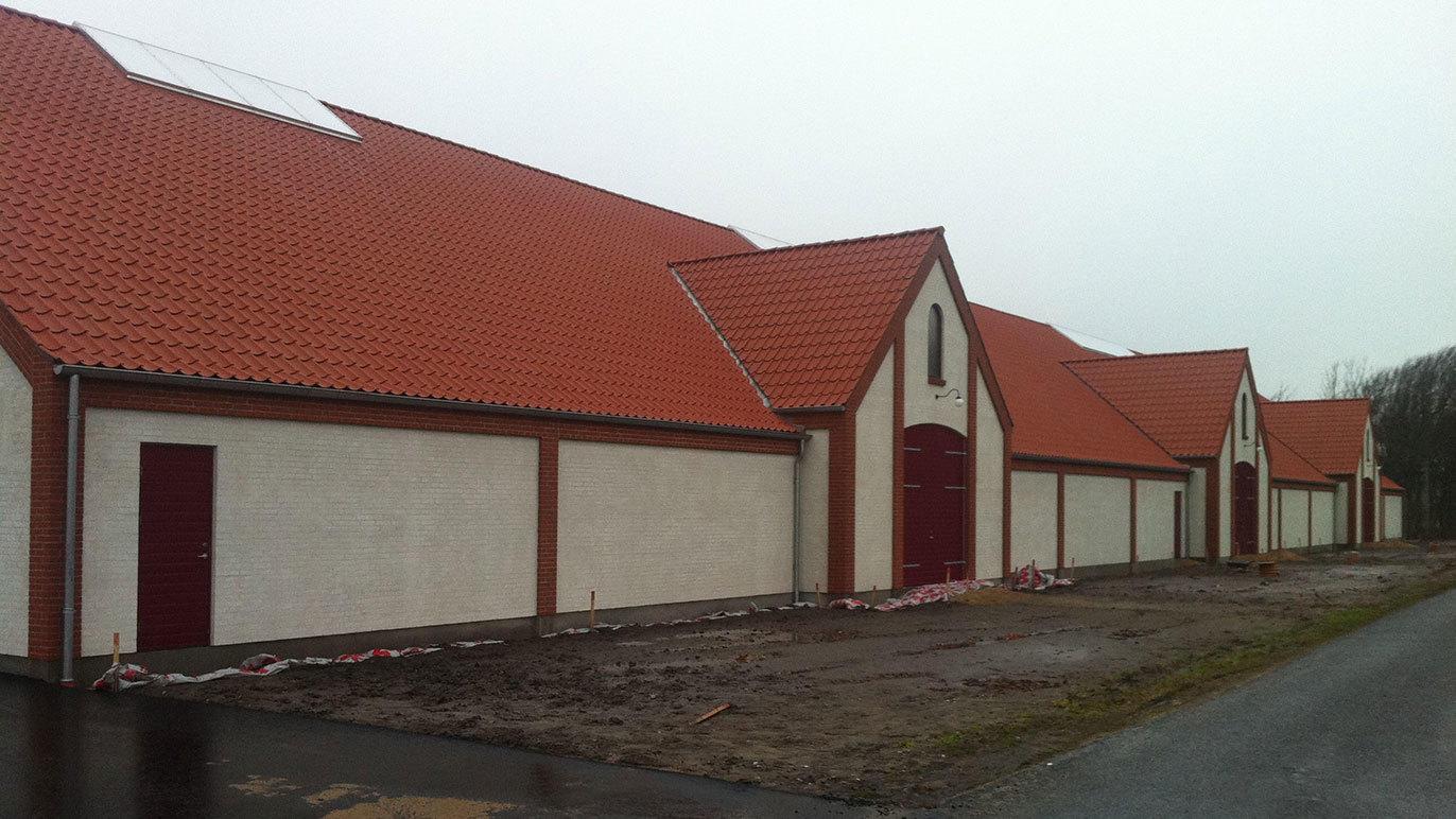 Række af bygninger med røde tage og hvide vægge langs en grusvej på en overskyet dag.