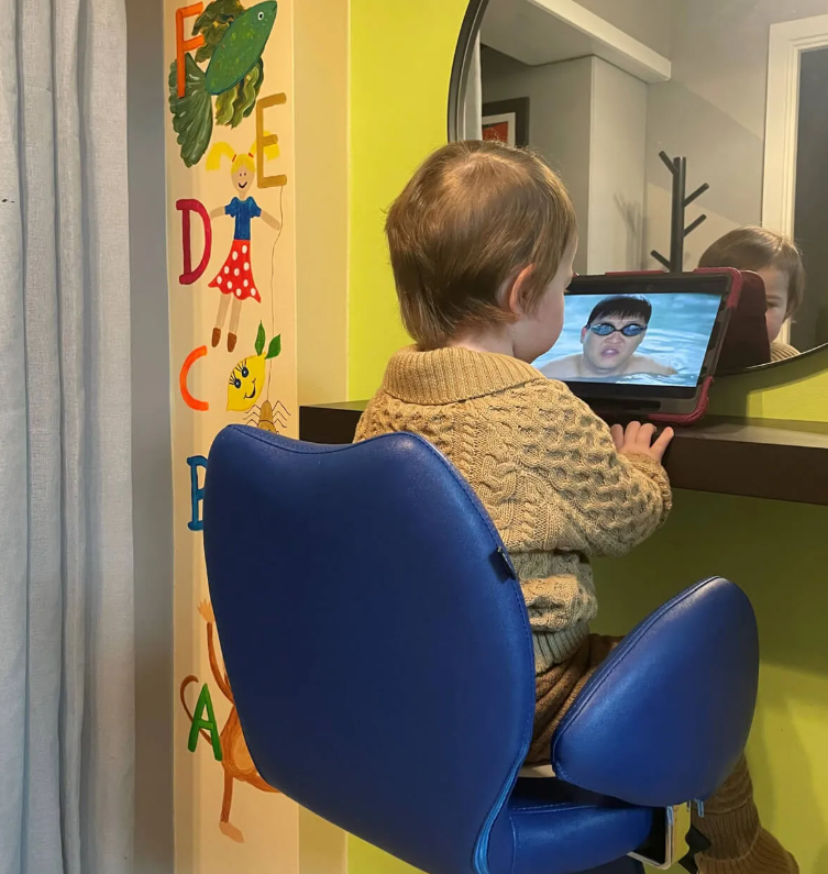 Barn sidder på en blå stol og ser en video på en tablet foran et spejl i et farverigt rum.