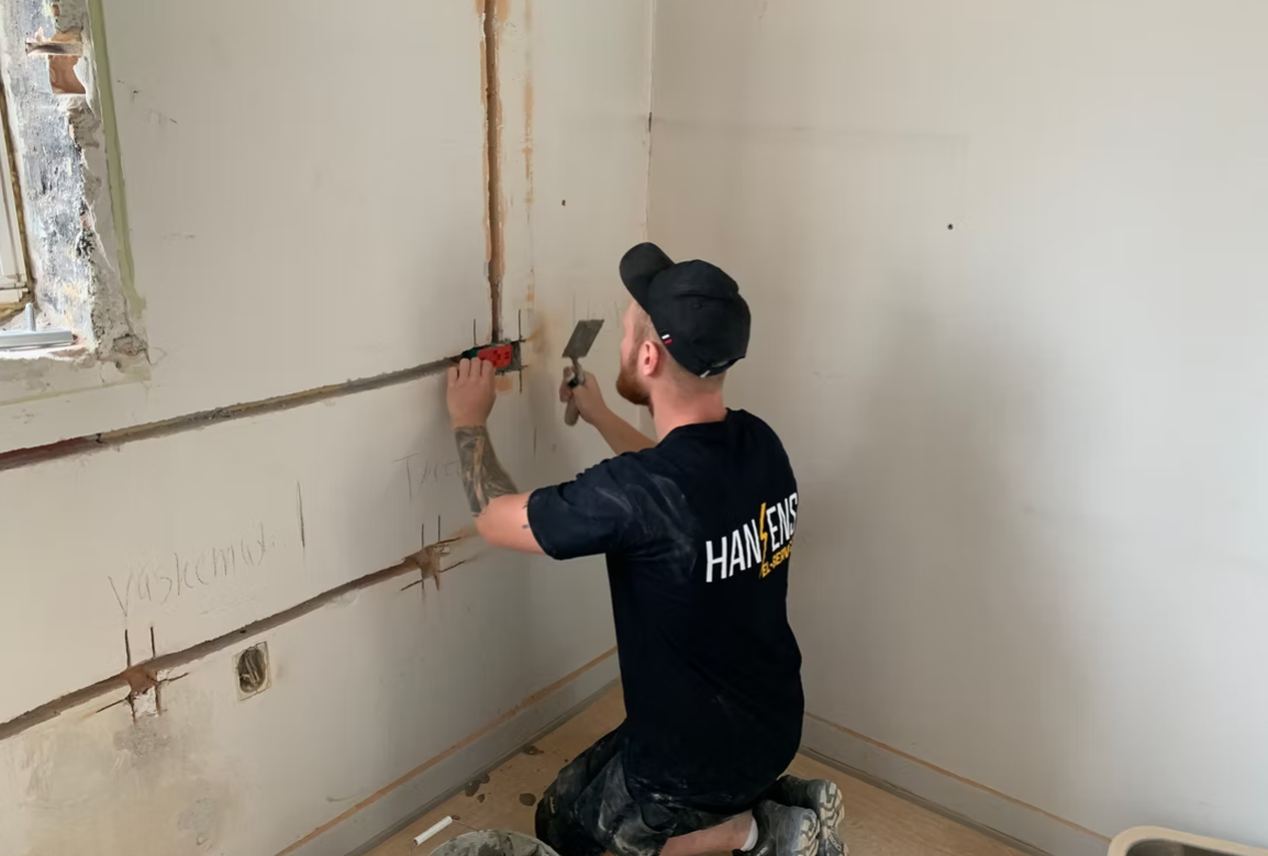 En håndværker, der står på knæ og arbejder med en spartel i et rum med ubehandlede vægge.
