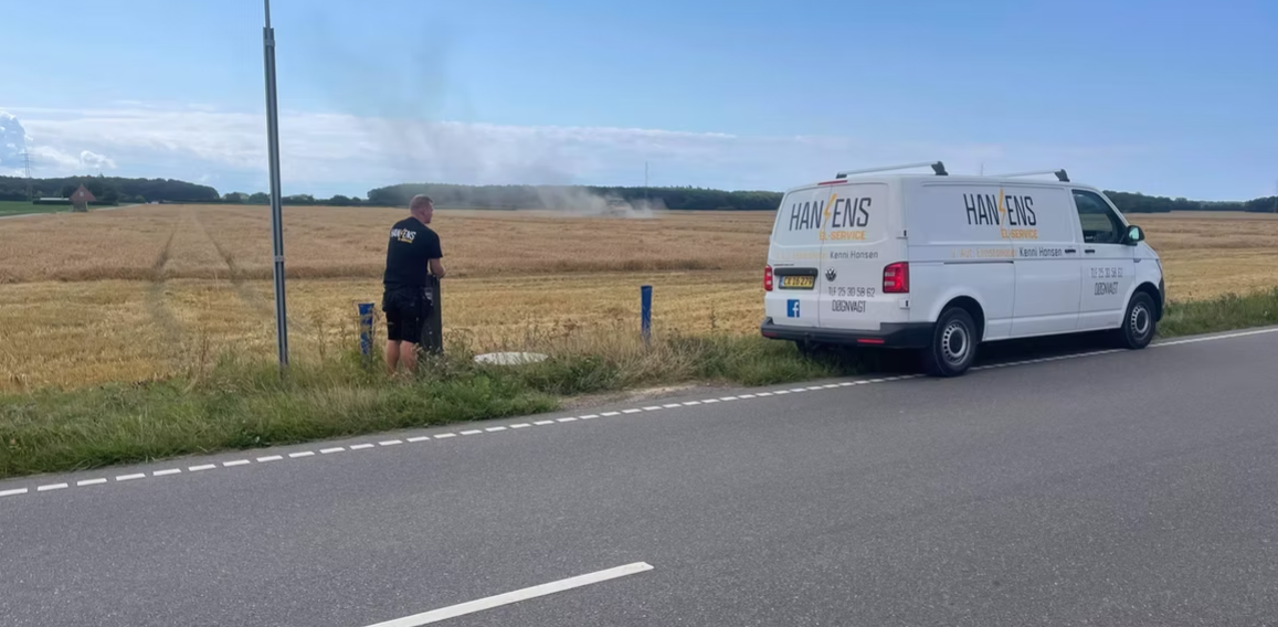 En mand står ved en mark med røg i baggrunden, mens en varevogn holder parkeret langs vejen.