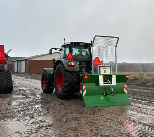 Traktor med en grøn sneslynge på en mudret vej ved en landbrugsbygning.