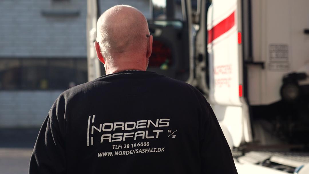 Mand iført sort T-shirt med teksten "Nordens Asfalt" på ryggen, står foran en lastbil.