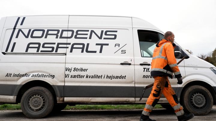 En arbejdstager i orange arbejdstøj går forbi en hvid varevogn med teksten "NORDENS ASFALT" på siden.