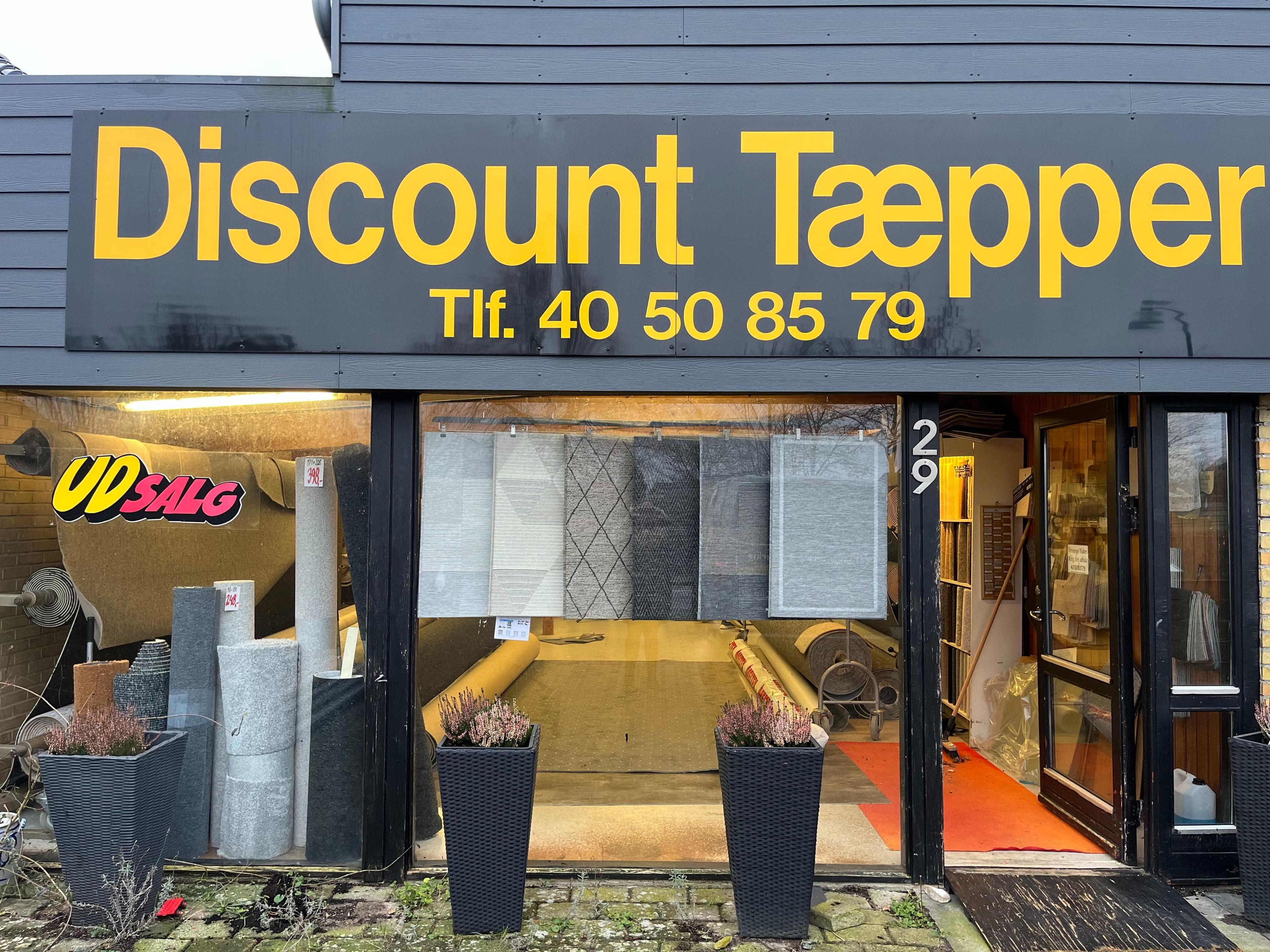 Butiksskilt for Discount Tæpper med telefonnummer og udstillingsvindue fyldt med tæpper.