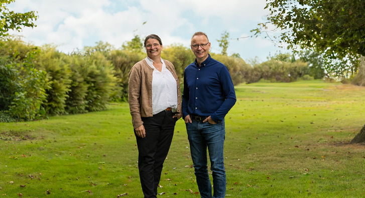 To personer står på en grøn græsplæne med træer i baggrunden, smilende og klædt i afslappet tøj.