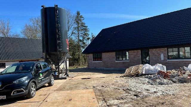 Billede af en byggetomt med en sort silo, en bil parkeret foran et hus og byggeaffald på jorden.