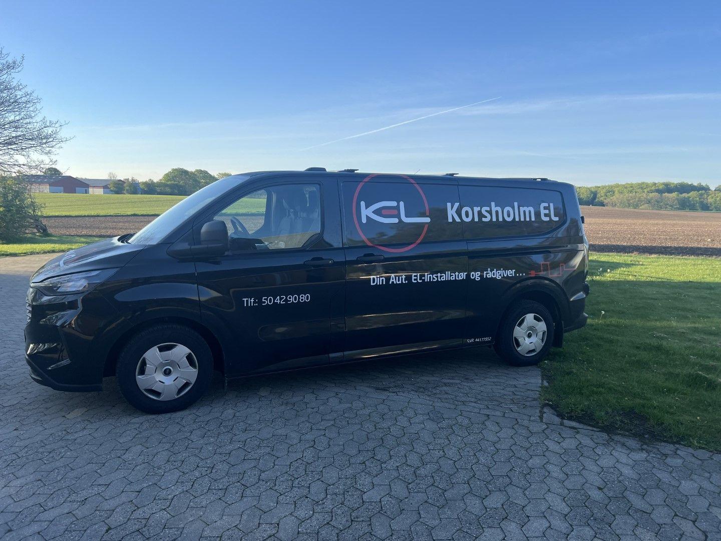 En sort varevogn med teksten "Korsholm EL" og kontaktinformation står parkeret på en grusvej.