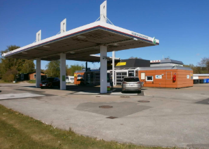 Benzinstation med flere pumper og en butik, omgivet af biler og grønne områder under en klar blå himmel.