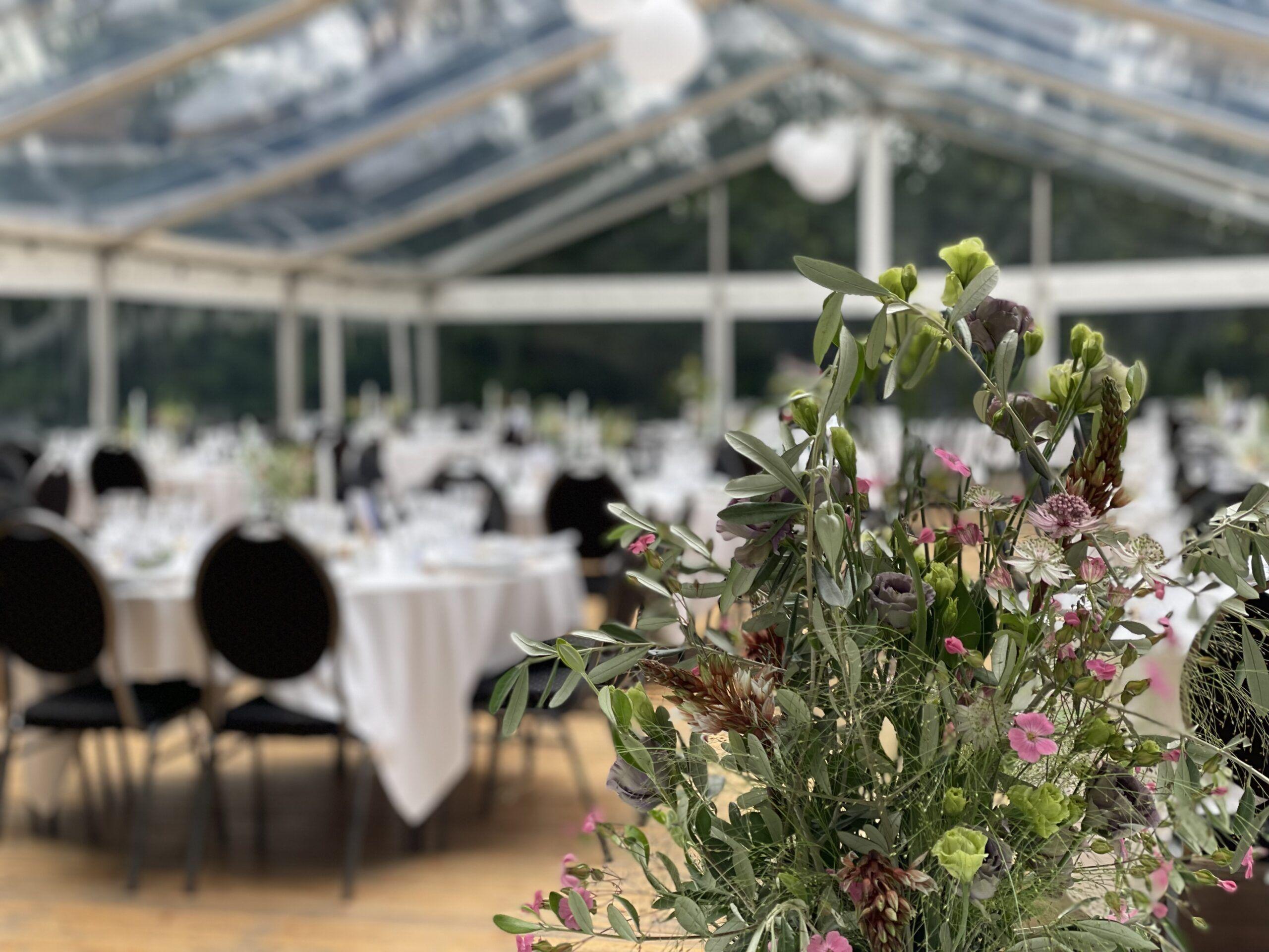 Blomsterarrangement i forgrunden med dækkede borde i en teltlokale til fest.