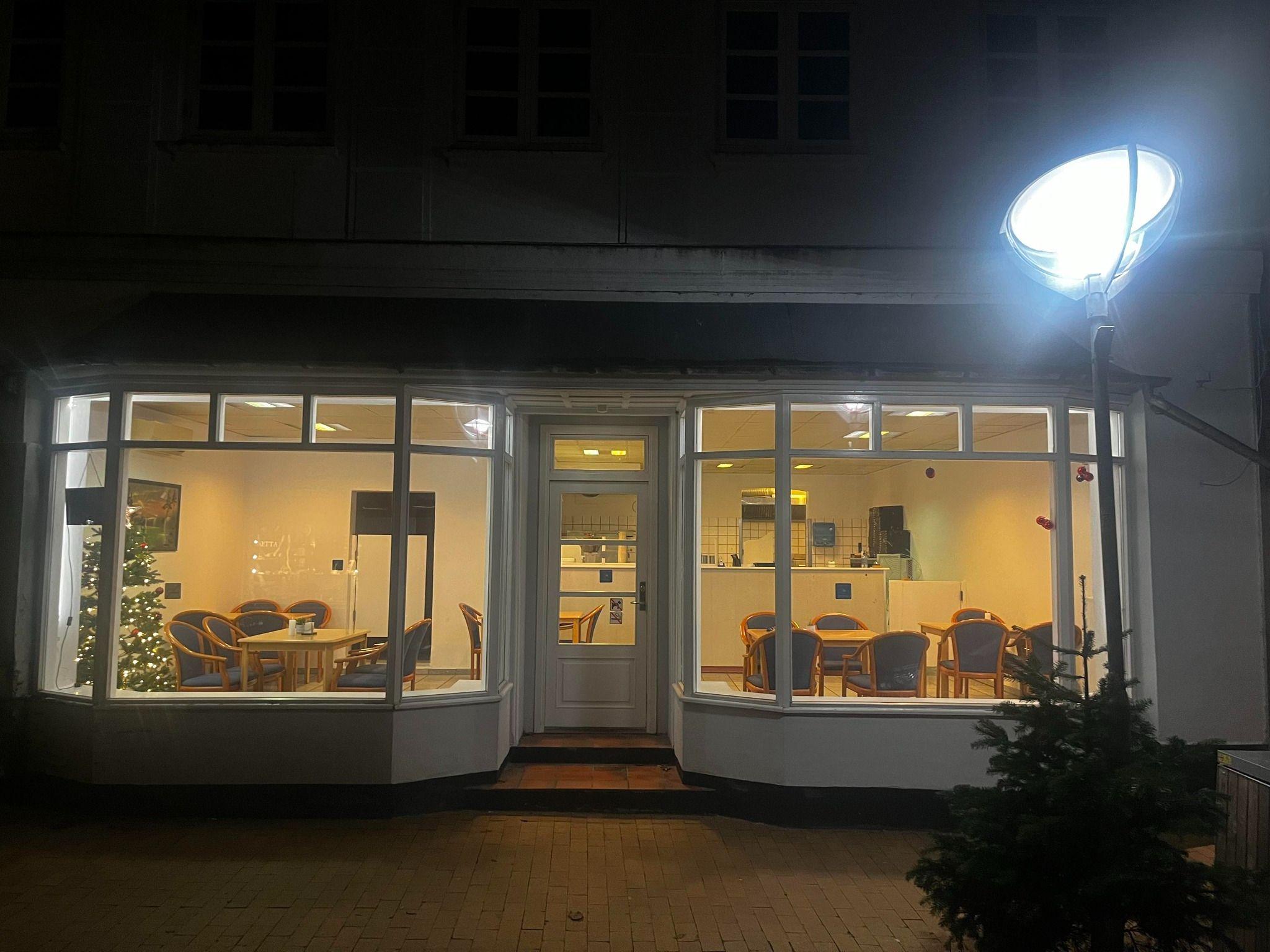 En café med store vinduer, der viser et lyst indendørsområde med borde og stole, oplyst af en lampe.
