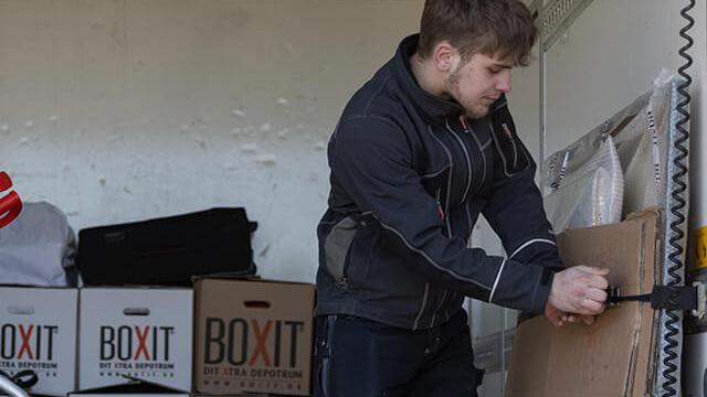 En ung mand låser en kasse i en flyttebil, omgivet af flyttekasser med logoet "BOXIT".