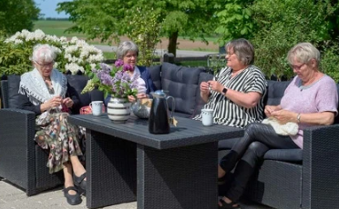 Fire kvinder sidder på en terrasse og strikker, omgivet af grønne planter og blomster.