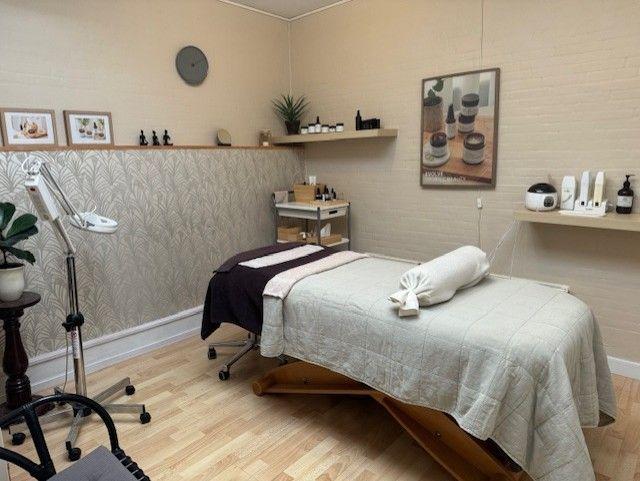 Et afslappende behandlingsrum med en massagebriks, hylder med skønhedsprodukter og en plante i hjørnet.