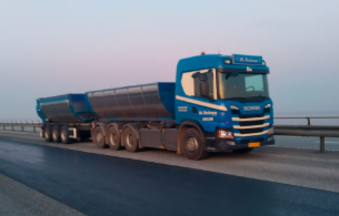 En blå Scania lastbil med to trailere kører på en bro ved solopgang.