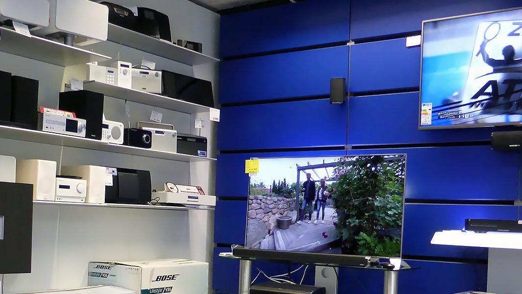 Fladskærms-tv viser en scene fra en udendørs aktivitet i en elektronikbutik med hylder fyldt med gadgets.