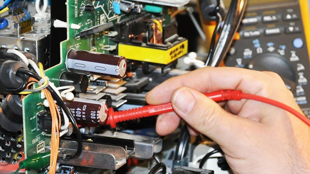 En hånd holder en rød multimeterprobe, der måler komponenter på et kredsløbskort i en elektronisk enhed.