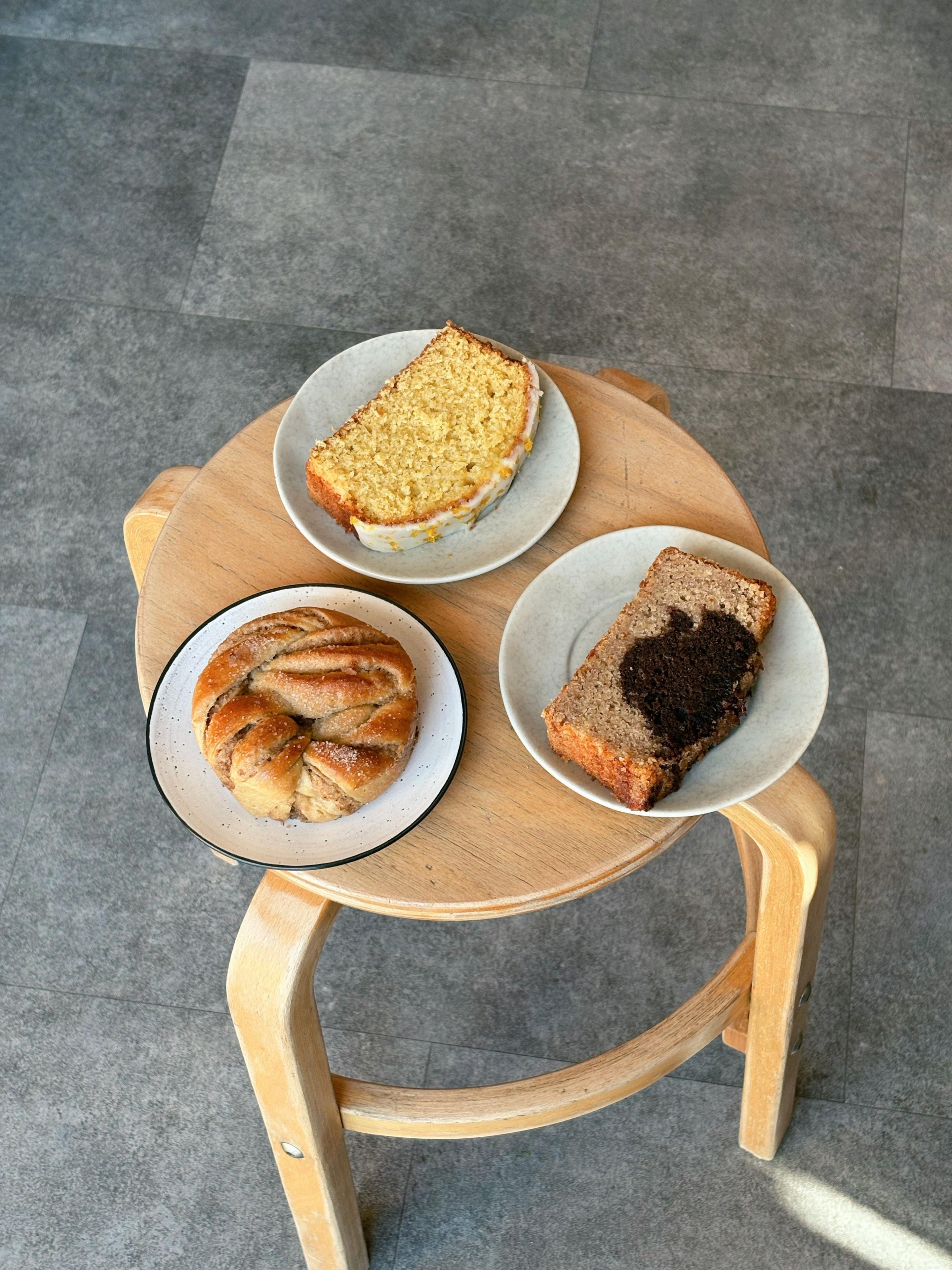 Tre tallerkener med bagværk: en kanelsnegl, en skive chokoladebrød og en skive kage på et lille træbord.