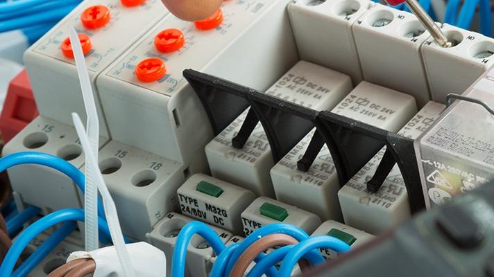 Nærbillede af elektriske relæer og ledninger i et kontrolpanel med farverige knapper og forbindelser.