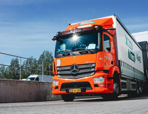 En orange lastbil fra Mercedes kører ind i en lastterminal under en klar blå himmel.