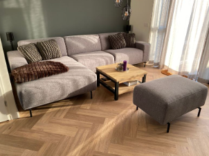 Moderne stue med en grå sofa, et træbord og en pouf, belyst af naturligt lys fra vinduet.