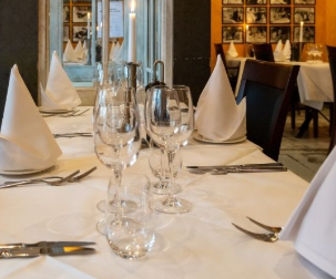 Dækket bord med hvide servietter, glas og bestik i en restaurant med spejl og billeder på væggen.
