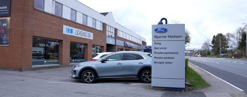 En sølvgrå bil parkeret foran en bygning med Ford-skilt og kontorvinduer.