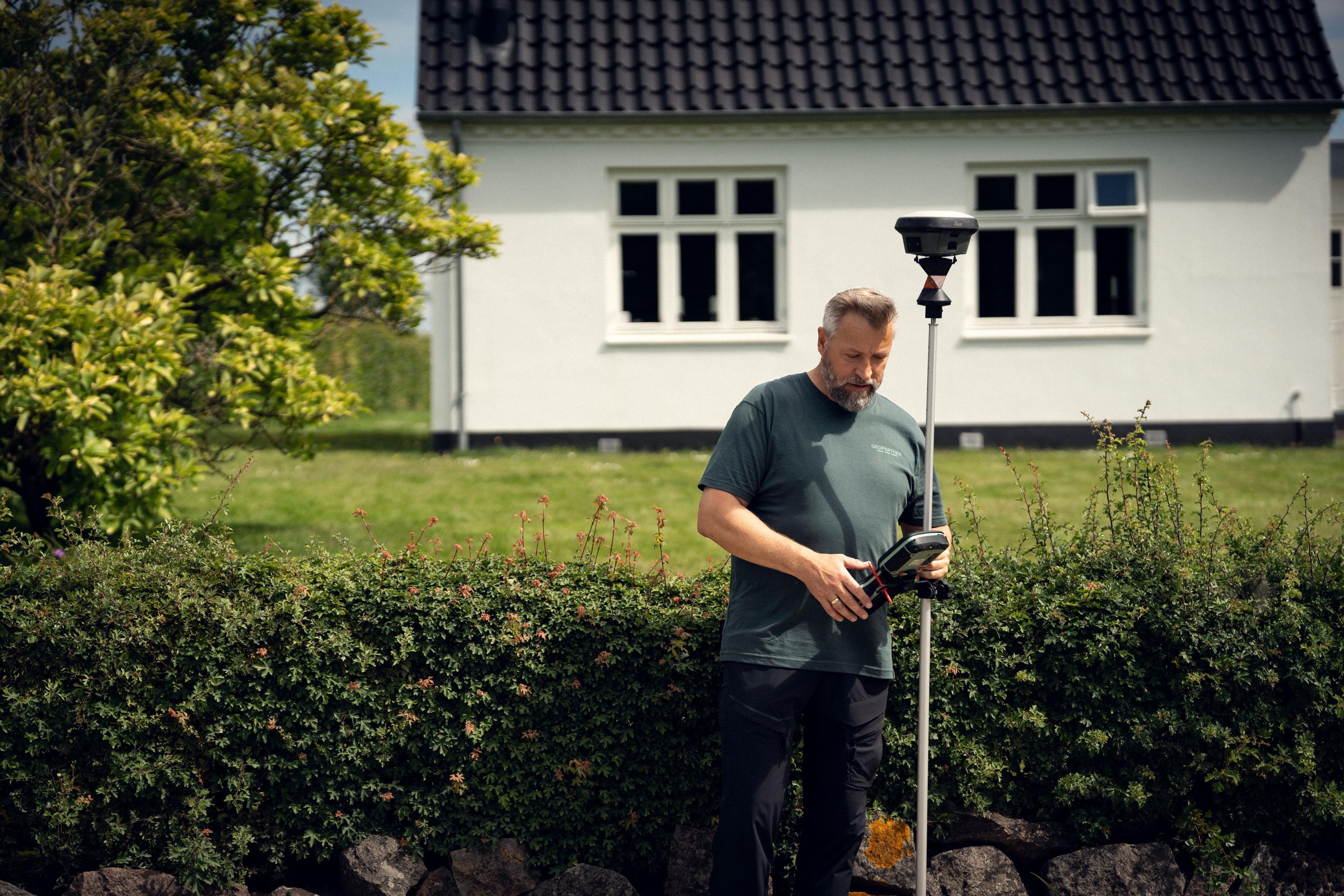 Mand står ved en hæk med en måleapparat i hånden, mens et hus ses i baggrunden.