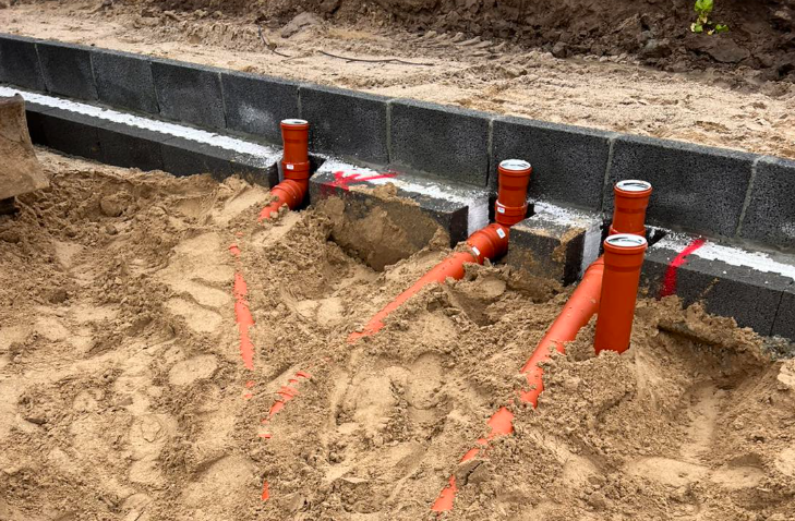 Rørinstallation i sandet ved en bygning, med orange rør og grå blokke i baggrunden.