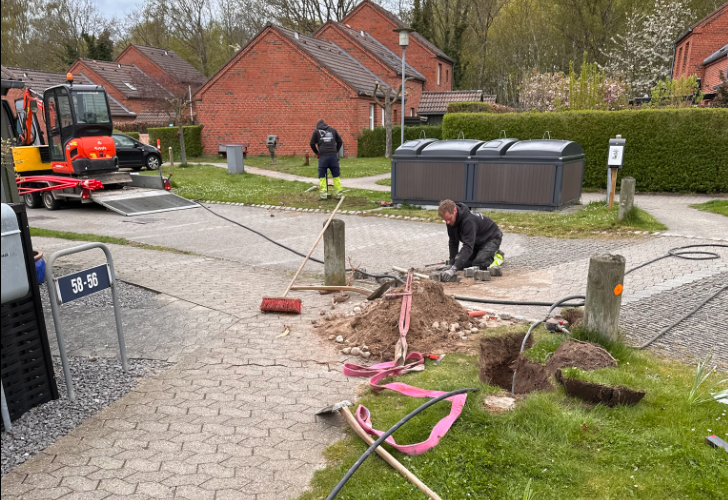 Arbejdsfolk graver i jorden på en boligvej, med redskaber og maskiner i baggrunden.