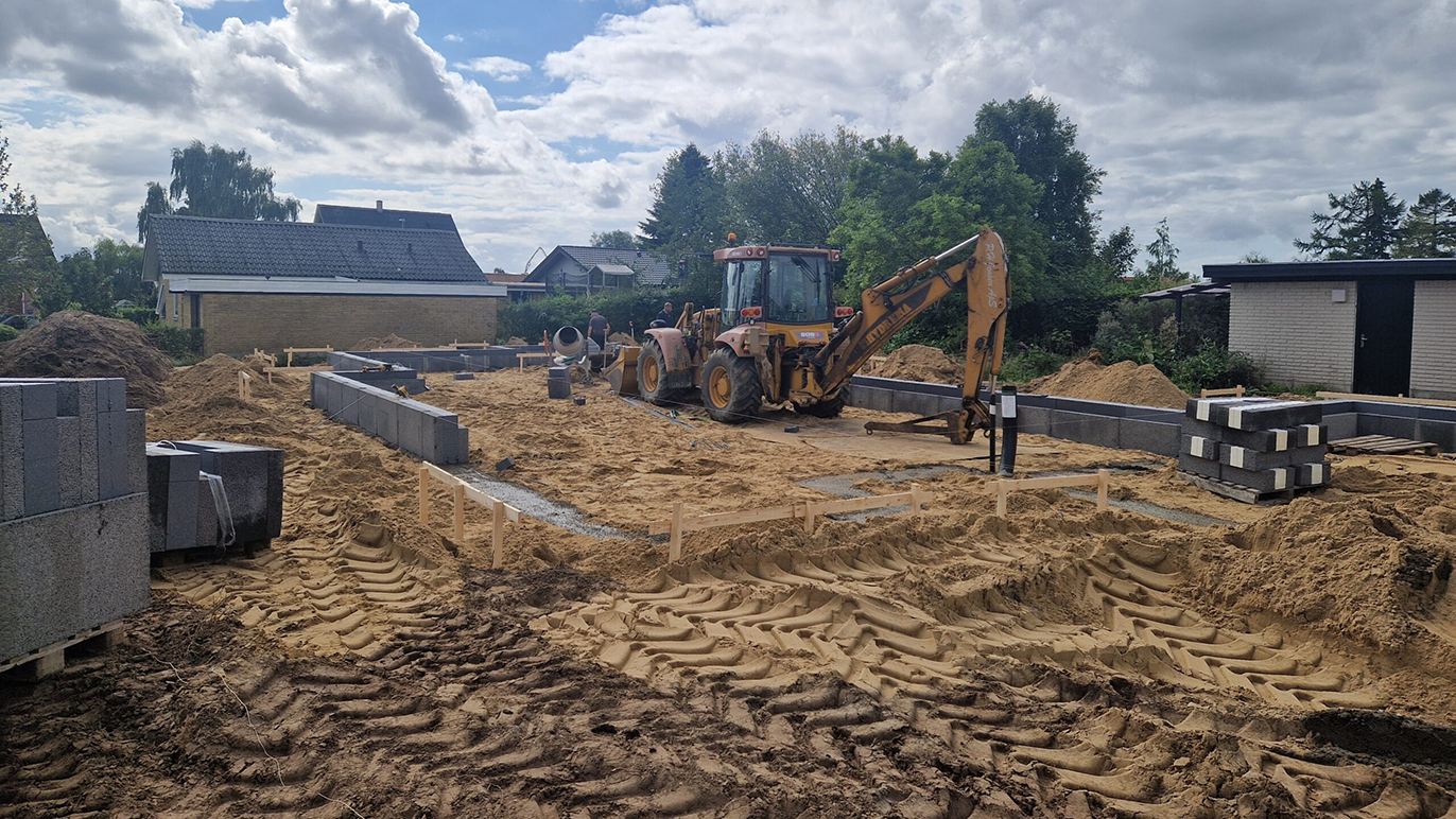Byggeplads med en gravemaskine, sand og fundamentblokke under konstruktion af et hus.